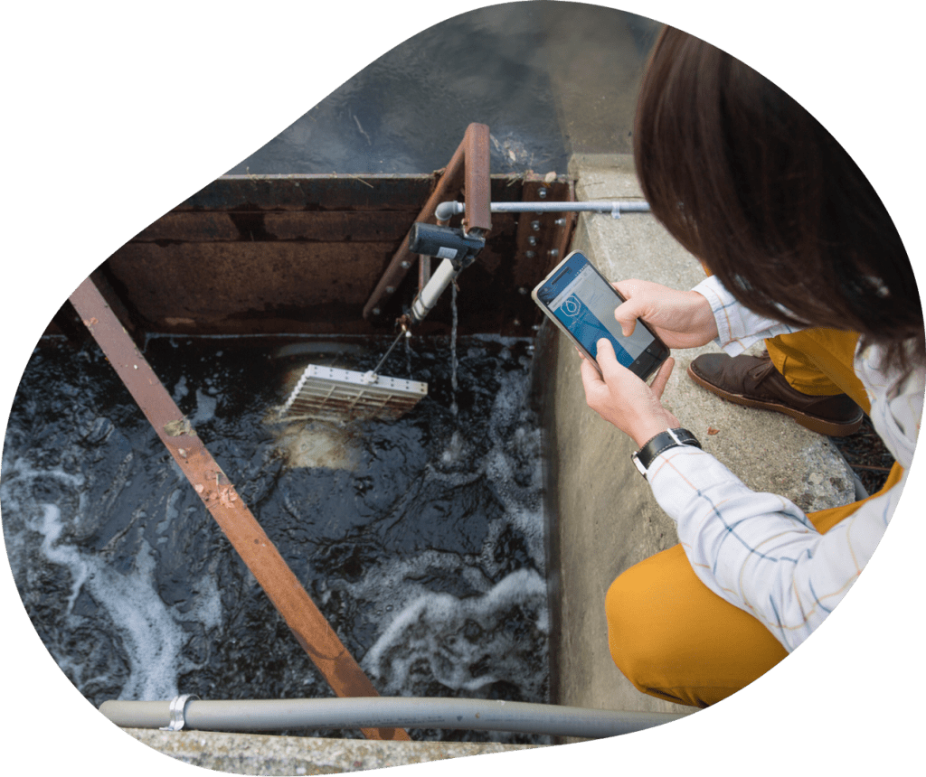 A person crouches next to a water dam with sensors and tubing attached to it. They are holding a cellphone with a water droplet pictured on the screen.