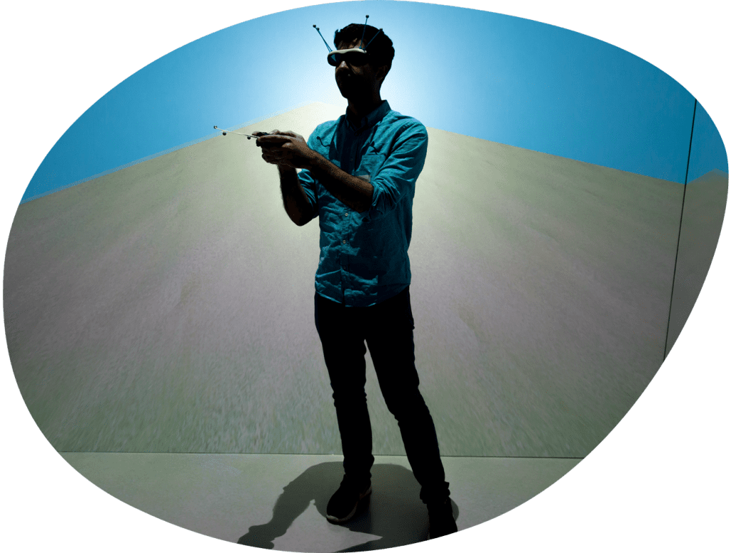 A person stands inside of a virtual reality platform, with screens on all sides showing what the person is seeing. The screens show a sandy terrain with a blue sky. The person is holding a controller and wearing goggles.