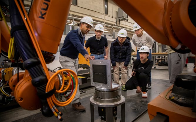 Five people in hard hats and safety goggles look towards a piece of equipment with a robotic arm in the foreground of the photo. One points to the piece of equipment.