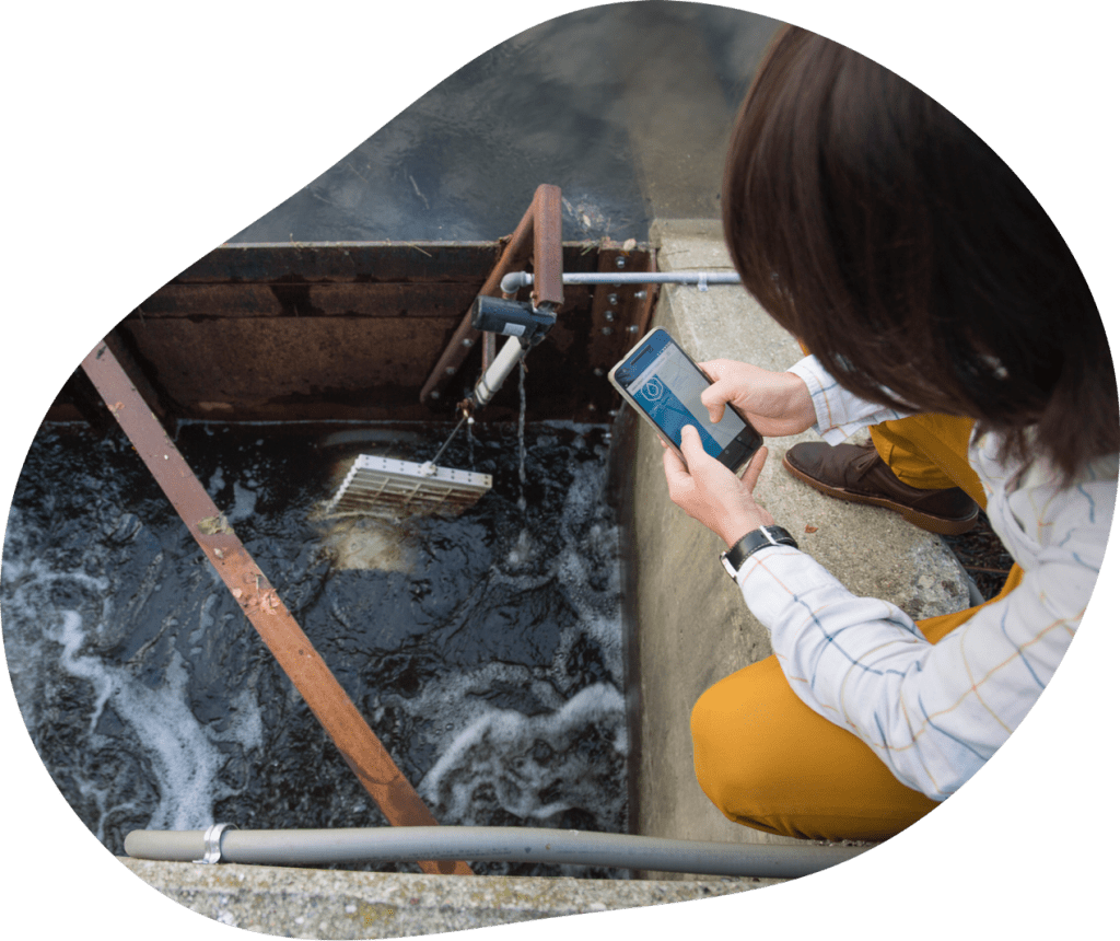 A person crouches next to a water dam with sensors and tubing attached to it. They are holding a cellphone with a water droplet pictured on the screen.