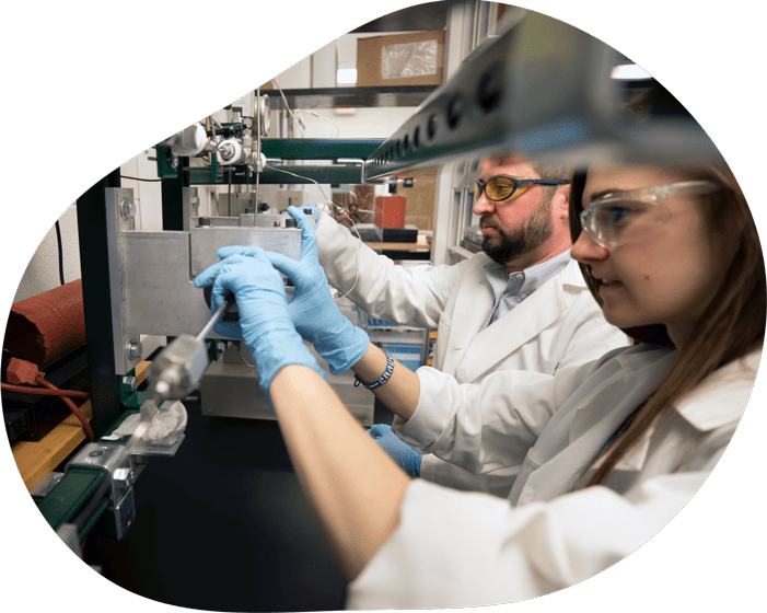 Two people in lab coats, safety glasses, and protective gloves work with instrumentation on a lab bench.