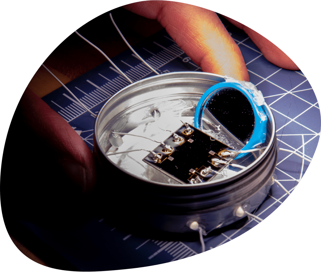 A person holds a small metal tin containing a small circuit board and a blue circular diode, with eight wires coming out of the tin at equally spaced points.