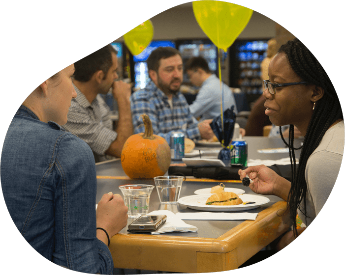 A group of people sit at tables together, eating and conversing. There are balloons and a pumpkin with "Go Blue" written on it all sitting on the tables.